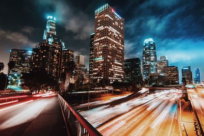 Light trails on road by illuminated buildings against cloudy sky at night