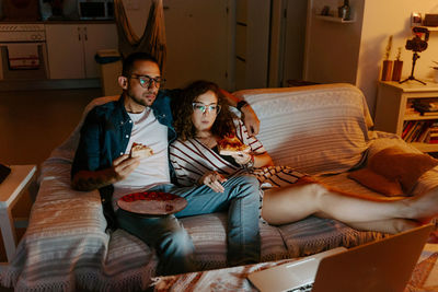 From above of young multiracial couple in casual clothes cuddling and eating pizza while sitting together on couch and watching movie on laptop