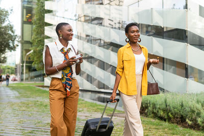 Optimistic young african american female tourists with short dark hair in stylish clothes smiling while walking on paved street with suitcase and photo camera in city