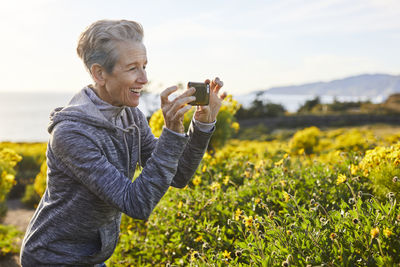 Cheerful senior woman photographing through smart phone while standing by plants against sky