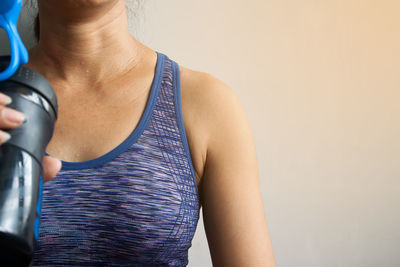 Midsection of mature woman holding water bottle while sitting against wall at home