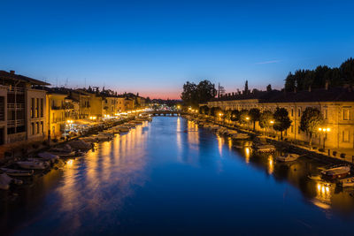 Boats moored on canal in illuminated city against clear blue sky