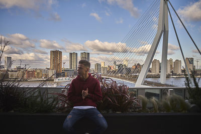 Man sitting on suspension bridge in city against sky