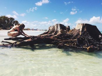 Side view of young woman wearing bikini while crouching on driftwood in sea against sky