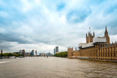 Thames river by houses of parliament in city against cloudy sky