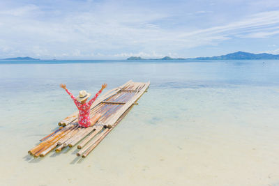 Woman with arms raised sitting on wooden raft at beach against sky