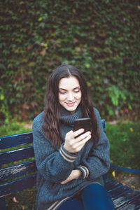 Young woman in a sweater holding a smart phone, sitting on the bench in the park. autumn time.