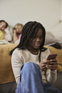 Female teenager using smart phone with friends lying in background at home