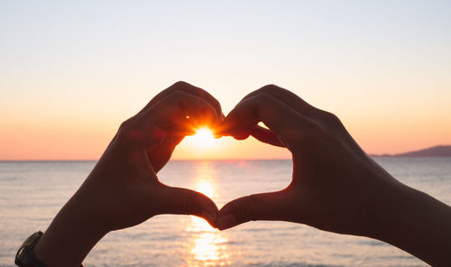 Close-up of hand holding heart shape against sky during sunset