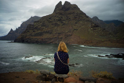 Rear view of woman sitting on rock by sea against mountains