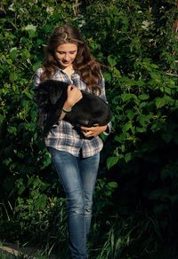 Young woman holding black labrador puppy against trees in lawn