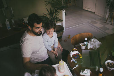 High angle view of father feeding sick son at table in living room