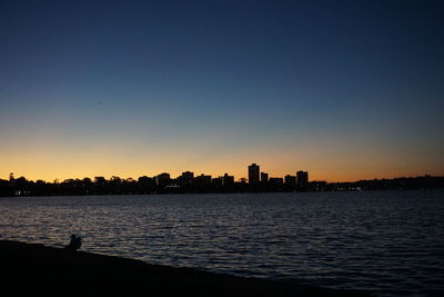 Silhouette buildings by sea against clear sky during sunset