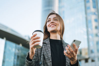 Young smiling woman in coat with coffee cup using mobile phone in evening city street