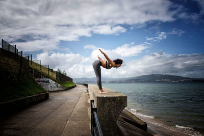 Side view of woman exercising by lake against cloudy sky