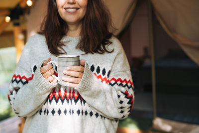 Brunette woman in nordic sweater drinking tea and relaxing in glamping in nature