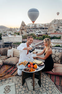 From above of cheerful male serving tea for wife while having romantic date on rooftop terrace against picturesque view of old city with hot air balloons flying