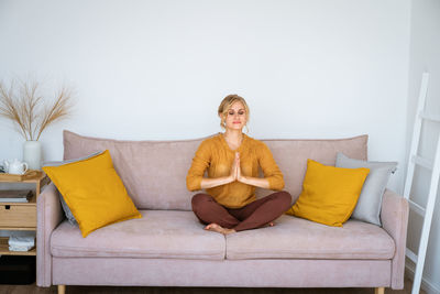 Young calm woman meditating in easy lotus position at home, sitting on sofa