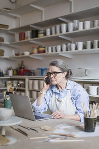 Senior female potter talking on mobile phone while using laptop at table in workshop