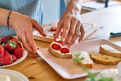 Female hands putting slices of ripe red strawberry on toast with spread stracchino cheese.