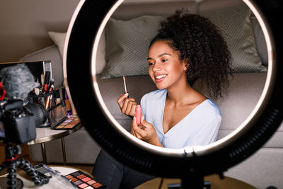 Smiling young woman applying make-up while being filmed in camera