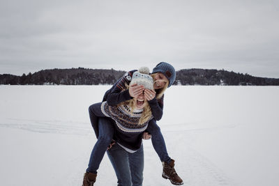 Playful woman covering friend's eyes while being piggybacked by her on snow during winter