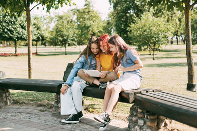 Three charming schoolgirls are sitting on a park bench and preparing for lessons or exams together 