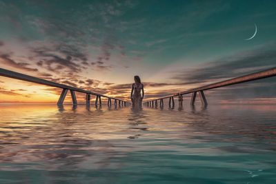 Woman standing amidst railing in sea against sky during sunset