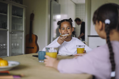 Girl eating bread slice while sitting at dining table with sister in home