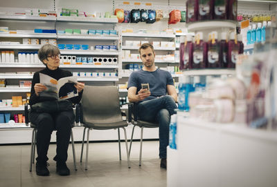 Customers sitting on chairs against medicine rack at pharmacy store