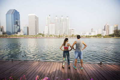 Rear view of couple standing with hands on hip at wooden walkway by river