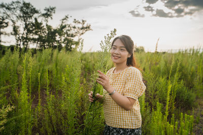 Smiling young woman holding while standing by plants against sky