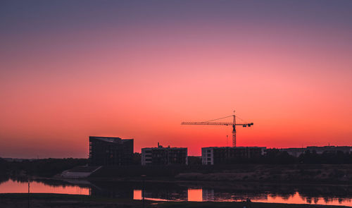 Reflection of crane and buildings in lake against sky during sunset