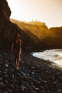 Woman standing on rock at beach against sky during sunset