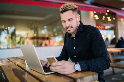 Young man using smart phone while working on laptop at restaurant