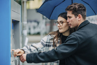 Young couple with umbrella using atm machine