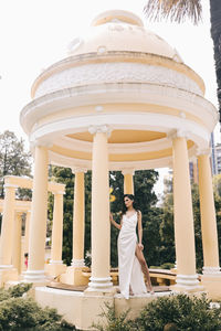 A beautiful brunette lady in an elegant wedding dress poses among the columns in the old city park