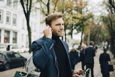 Businessman looking away while holding in-ear headphones in city