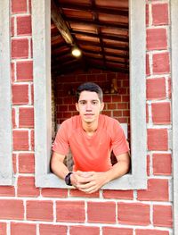 Portrait of young man standing at window amidst brick wall