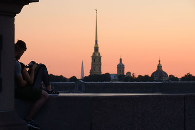 Couple sitting on railing against clear sky during sunset