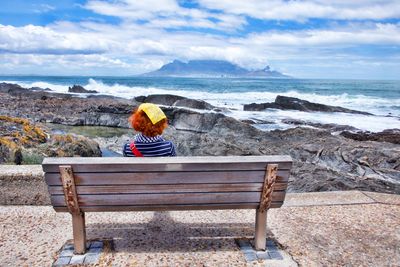 Rear view of mature woman looking at sea while sitting on bench against cloudy sky