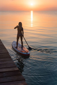 Back view of silhouette of unrecognizable female surfer standing on paddleboard and rowing against spectacular sun in sunset sky