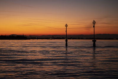 Silhouette street light by river against sky during sunset