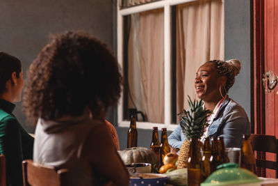 Cheerful diverse friends talking and smiling while sitting at table with beer bottles and vegetables sitting on terrace at night and having party