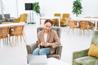 Adult smiling brunette business woman in beige suit and jeans working on laptop at public place