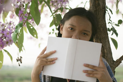 Portrait of girl holding book against tree