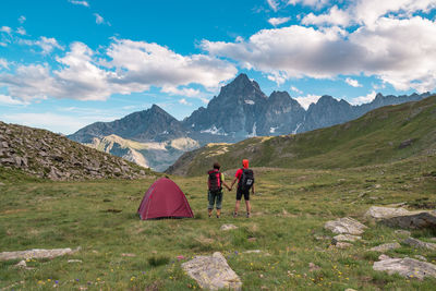 Rear view of backpackers on field against mountains and cloudy sky