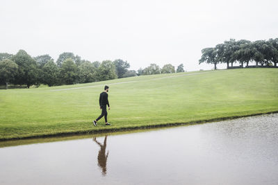 Young man walking by river on grassy land against sky