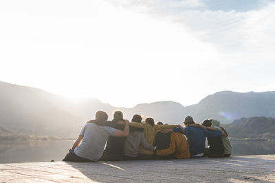 Male and female friends sitting on promenade with arms around during sunny day