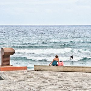 Rear view of woman sitting on retaining wall while man surfboarding in sea